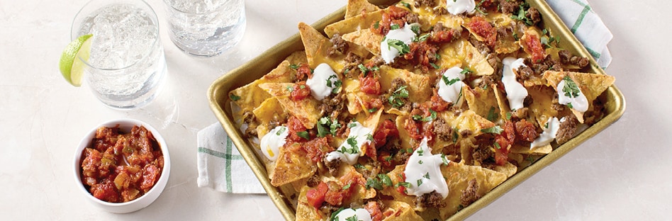 Sheet pan of nachos topped with seasoned beef, shredded cheese, sour cream and salsa.