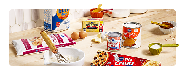 A whisk in a mixing bowl on a counter with other baking tools and ingredients such as sugar, flour, butter and canned goods.