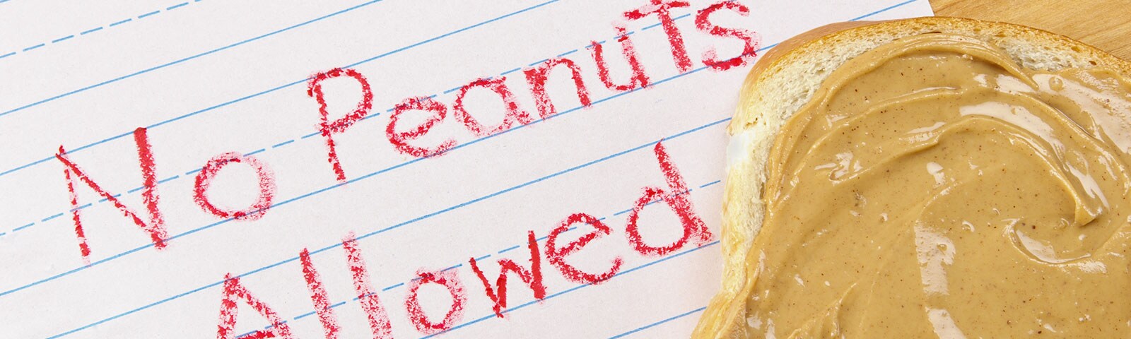 No Peanuts Allowed written on a piece of paper in crayon with a piece of bread slathered in peanut butter sitting on top of it