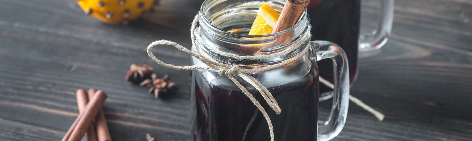 On a rustic wooden table two mason jars sit, wrapped in twine and filled with a dark beverage, an orange slice, and a cinnamon stick On a rustic wooden table two mason jars sit, wrapped in twine and filled with a dark beverage, an orange slice, and a cinnamon stick