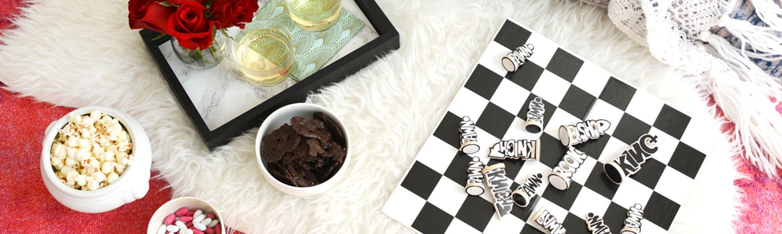 Atop a white shag rug sits a bowl of popcorn, a tray with roses and glasses of wine, a bowl of chocolate and a chess board.