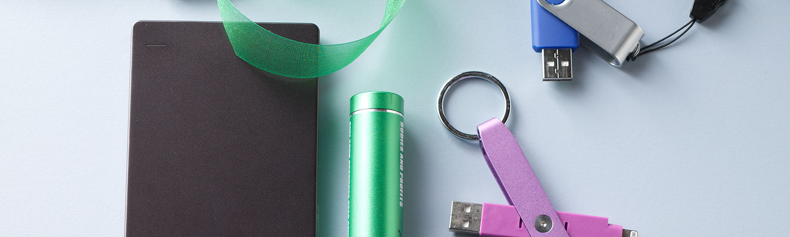 White table top with a flash drive, external hard drive, earbud headphones, spiral notebook and USB cord scattered about
