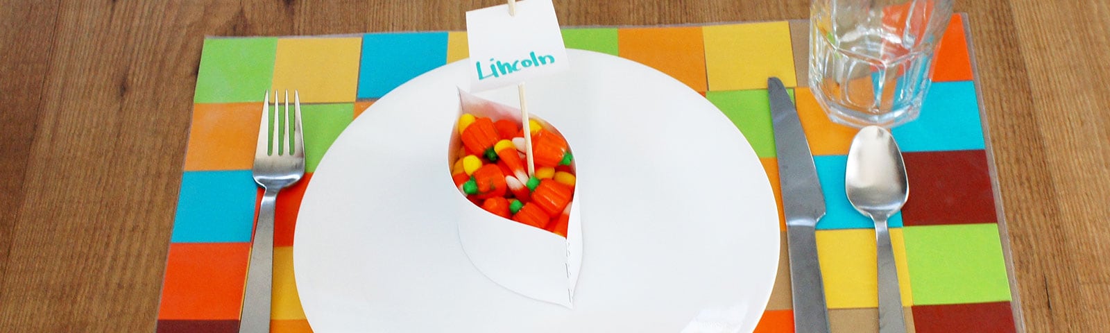 A colorful grid placemat on a wooden table with a white plate and a small jar filled with candy corn on top