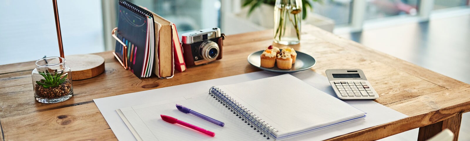 Pen and paper on wooden desk with cupcakes and plant