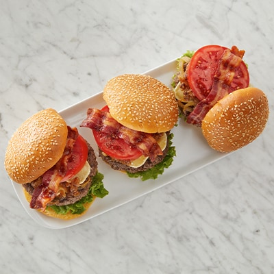 Three savory burgers with bacon, tomato, cheddar cheese, fried onions, and lettuce on sesame seed buns.